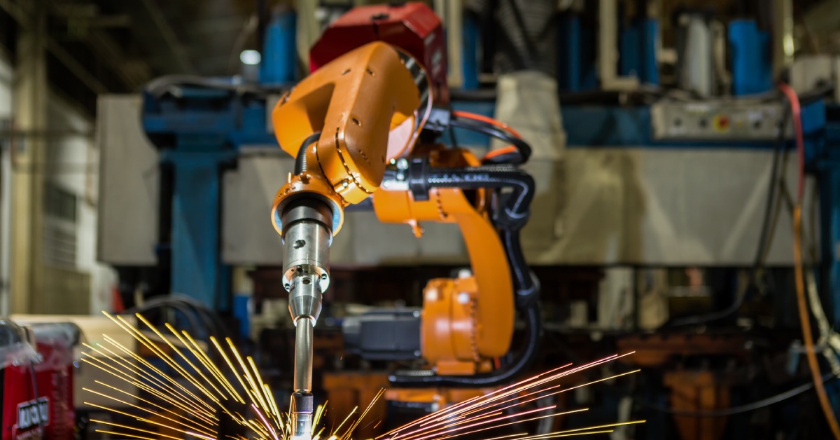 An orange automated industrial robot is welding automotive parts in a car factory. Sparks are flying around it.