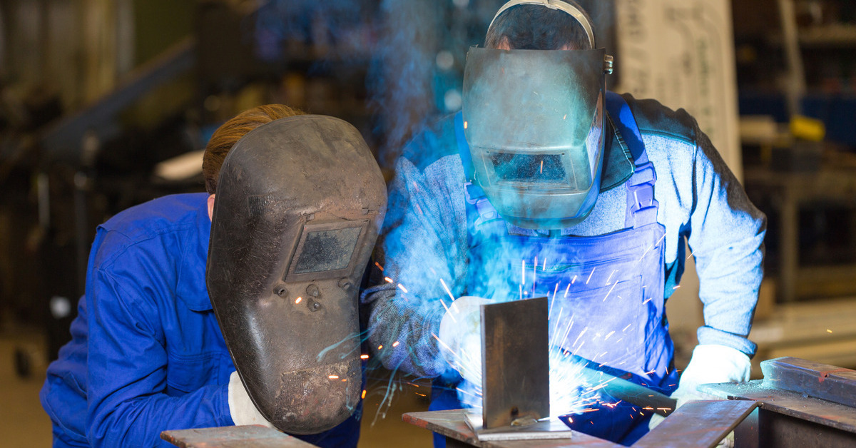 Two steel construction workers wearing protective gear and welding metal beams, with sparks flying in different directions.