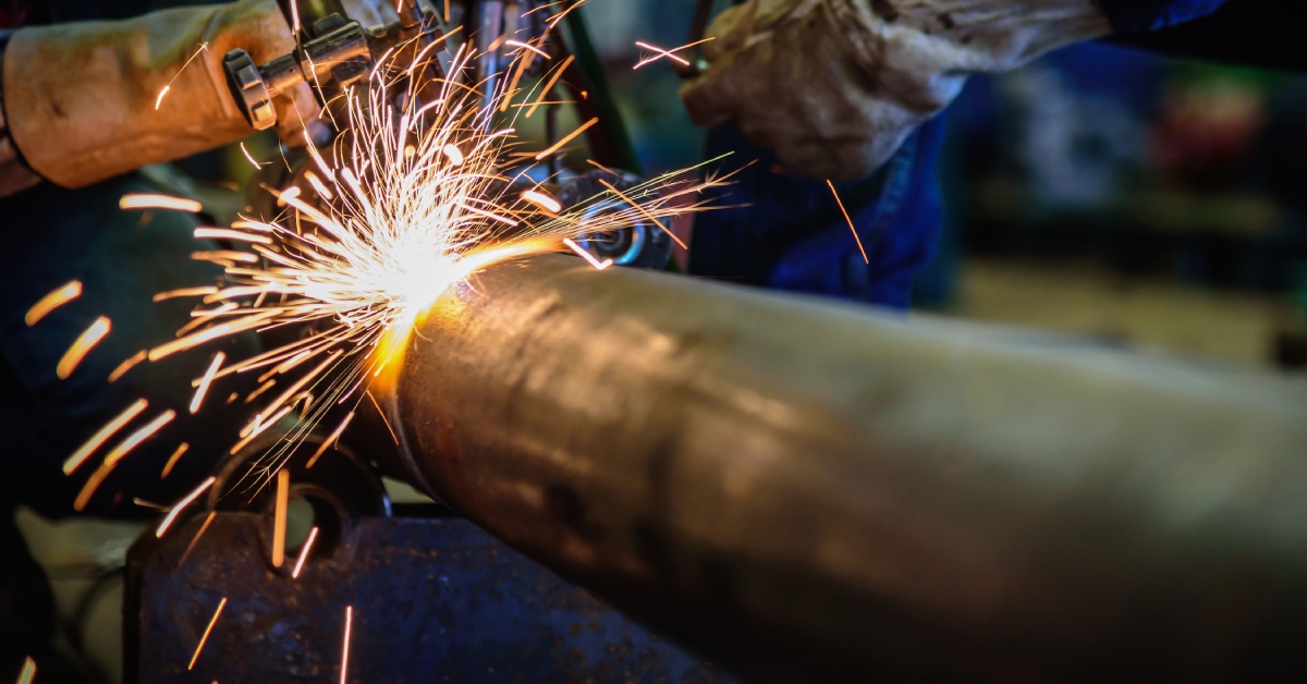 A welder works on a large steel pipe with equipment that sends sparks flying. He seems to be cutting or welding that section of the pipe.