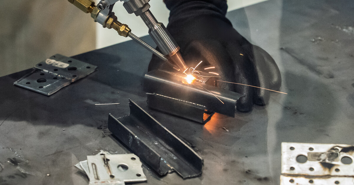 A professional welder uses a welding head for precise work on a small metal piece on a metal surface.