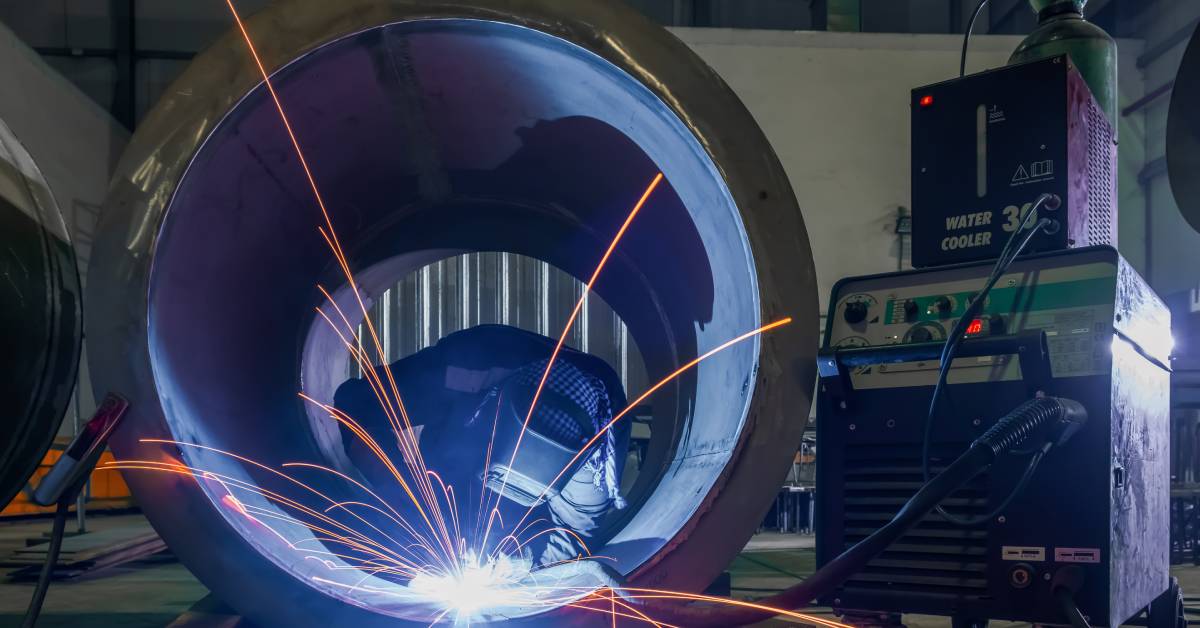 A welder with safety gear crouches inside a large metal pipe, performing a weld using a machine placed next to them.