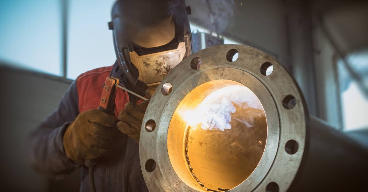 A welder in a protective mask and gloves carefully welds a steel pipe. Smoke emits at the contact point.