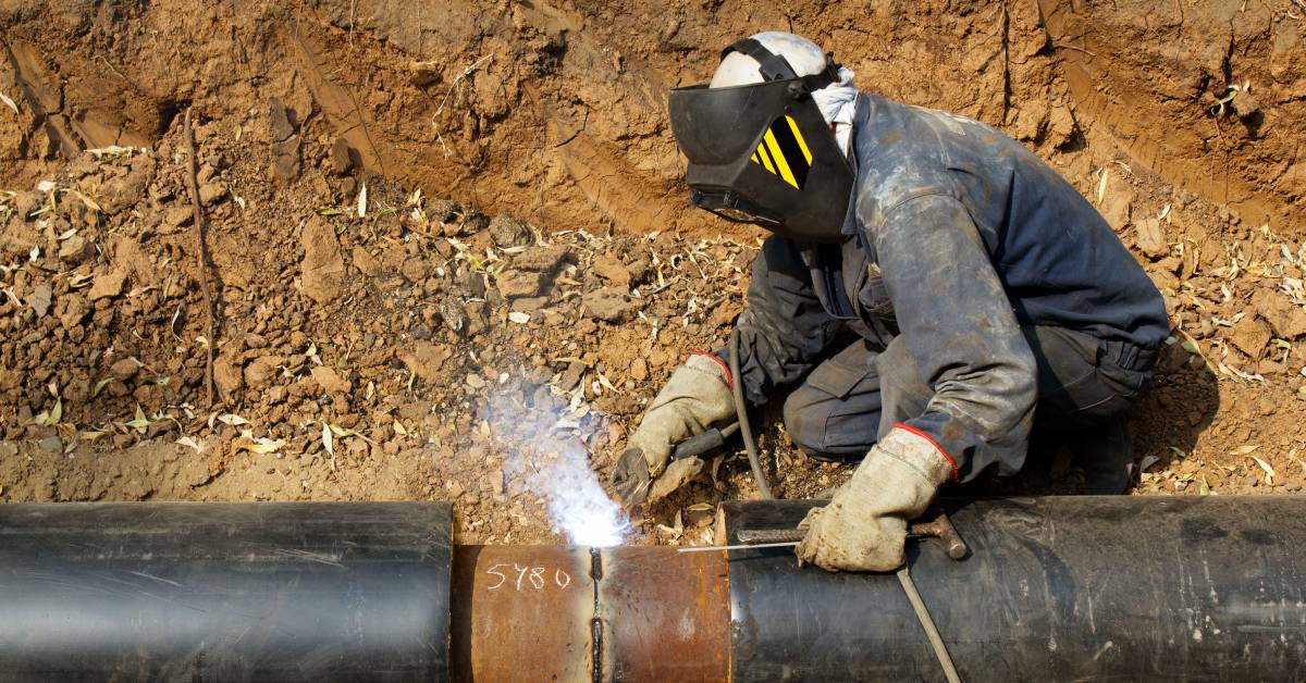 A welder works on a large metal pipe to secure the joint. They wear a mask and gloves for safety.