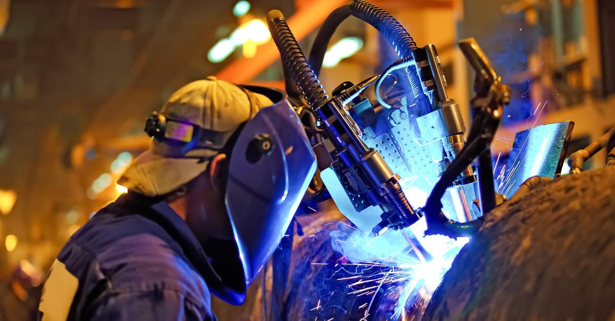 A welder stands close to an automated welding system on a large metal pipe. Blue light and sparks emanate from the pipe.