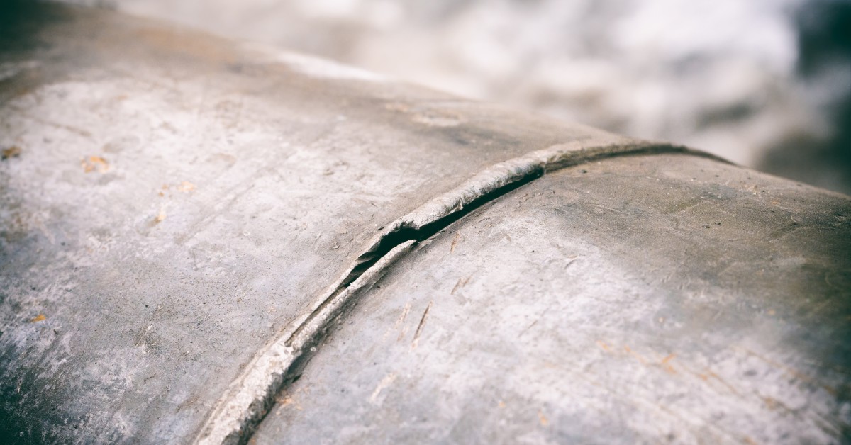 A close-up of a steel pipeline shows a visible crack along the welded seam, with the background softly blurred.