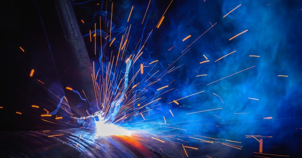 Sparks fly from a contact tip as it welds a round steel part in a dark space, lit only by the bright flashes.