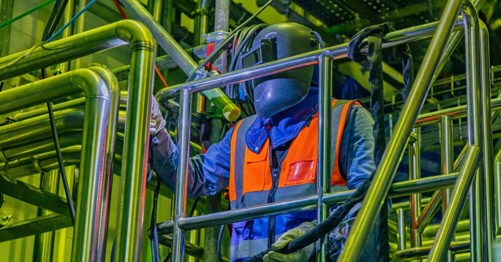 A person in a welding mask, gloves, and orange vest holds black cabling while inspecting metal piping in a tight space.