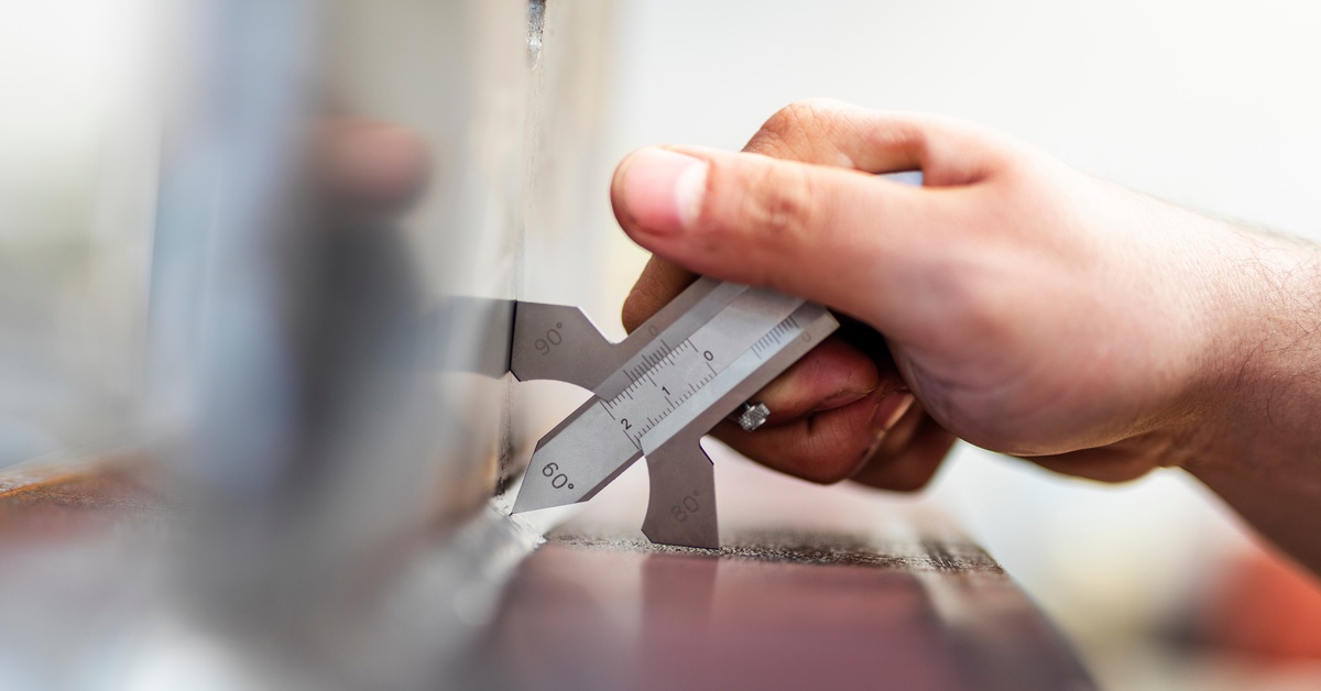 A hand holds a fillet gauge against a bent metal surface to measure the metal's thickness. The metal and background are blurred.