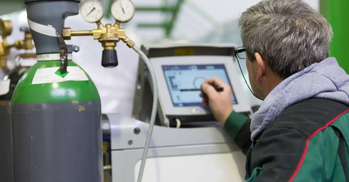The back of a man using a stylus on a touchscreen next to a gas canister with two gauges and a tube.