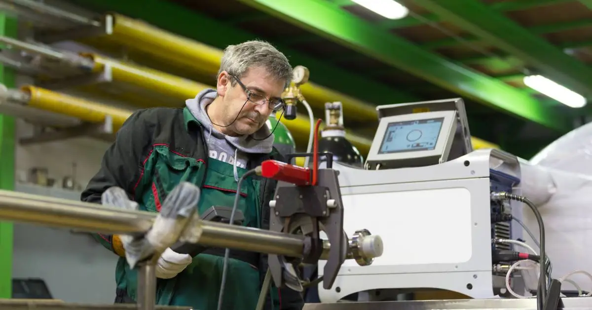 A man in glasses holds a black device and looks at an orbital welding machine with a pipe positioned inside it.